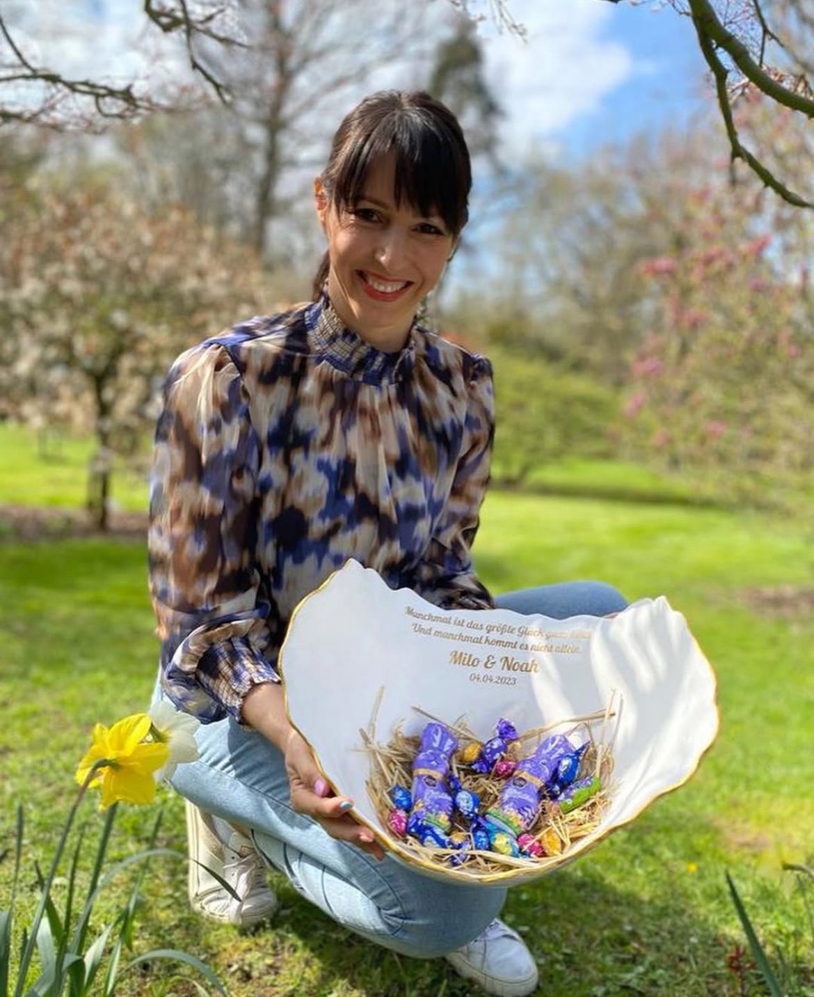 Eine kniende Frau in einem sommerlichen Garten, die eine Babyschale aus Gips mit Oster-Süssigkeiten präsentiert.