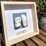 Gips-Abdrücke von Hand und Fuß eines Neugeborenen auf einem blauen Hintergrund mit Strich-Muster in einem hellbraunen Bilderrahmen mit Passepartout, der auf einem Holztisch neben einer Pflanze steht.