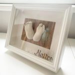 3D-Abdrücke aus Gips von den Füßen eines neugeborenen Babys auf einem Hintergrund mit Holzoptik in einem weissen Bilderrahmen mit Passepartout und dem Namen des Kindes.