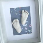 Gips-Abdrücke von den Füßen eines neugeborenen Babys auf einem blauen Hintergrund mit bunten Schmetterlingen in einem weissen Bilderrahmen mit Passepartout.