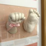 3D-Abdrücke aus Gips von der Hand und dem Fuß eines Neugeborenen Babys in einem braunen Bilderrahmen mit rosa Hintergrund und Punkten in weissem Passepartout.