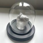 Ein 3D-Abdruck aus Gips von der Hand eines neugeborenen Babys in einer Glasglocke mit schwarzem Sockel.