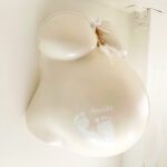 Ein 3D-Gipsabdruck eines Babybauches von Brust und Bauch der Schwangeren, gestaltet in der Farbe Beige mit weisser Banderole, Schleife, Federschmuck und dem Namen und Fußabdruck des Babys auf dem Bauch.