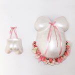 Der Abdruck eines Babybauches von Brust und Bauch aus Gips mit Blumen an der Unterseite neben dem Gipsabdruck eines Babypopos. Beide gestaltet mit rosa Bändern und in den Farben Weiss und Perlmutt.