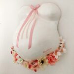Ein Babybauch-Abdruck einer schwangeren aus Gips mit rosa Schleife und Banderole, Kunstblumen an der Unterseite und dem Namen und Geburtsdatum des Kindes.