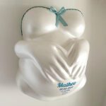 Der weisse Gipsabdruck eines Babybauches von Brust, Bauch und Händen die den Bauch umschlingen, gestaltet mit einer blauen Banderole und Schleife und dem Namen, dem Geburtsdatum und Geburtszeit, Geburtsgewicht und Geburtszeit des Kindes.