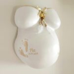 Ein weisser 3D-Gipsabdruck eines Babybauches von Brust und Bauch, gestaltet mit einer Banderole und Schleife in Ocker-Farbe und Federschmuck und den Fußabdrücken, Namen und Geburtsdatum des Babys.