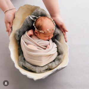 Ein neugeborenes Baby mit einem Blumenkranz auf dem Kopf, das in eine Decke eingekuschelt in einer Babyschale aus Gips liegt, präsentiert von den 2 Händen der Mutter.