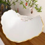 Eine Bellybowl oder Babyschale aus Gips in Weiss und Gold mit den Namen und Geburtsdaten der Neugeborenen in goldener Beschriftung. Im Hintergrund ist ein Osterstrauch.