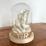 Der Gipsabdruck einer Menschenhand, die eine Hundepfote hält in der Farbe Gold, davor der Name des Hundes in Lasercut-Buchstaben. Beides präsentiert in einer Glocke aus Glas mit einem Sockel aus hellem Holz stehend auf einem Holz-Tisch.