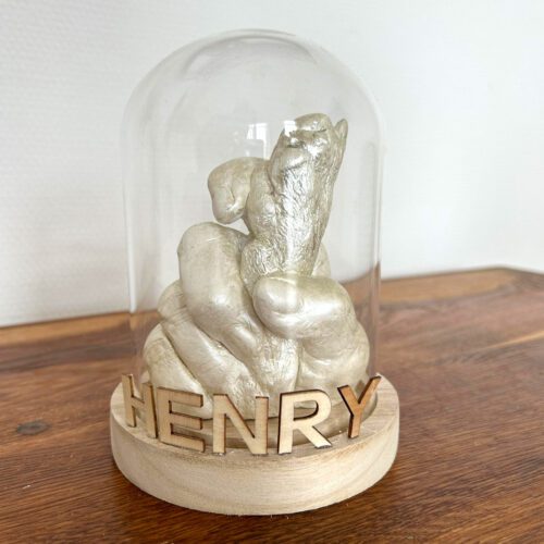 Der Gipsabdruck einer Menschenhand, die eine Hundepfote hält in der Farbe Gold, davor der Name des Hundes in Lasercut-Buchstaben. Beides präsentiert in einer Glocke aus Glas mit einem Sockel aus hellem Holz stehend auf einem Holz-Tisch.